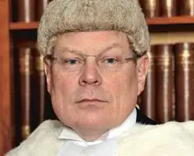 Man wearing a courtroom wig with legal books in background.