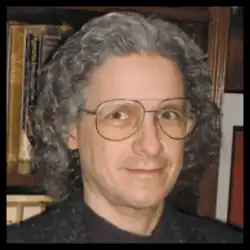 Portrait of Jeffrey Owen Katz, an American scientist with curly hair and glasses, standing in front of a bookshelf.