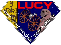 A diamond-shaped crest houses artworks of the Lucy fossil at left, the Lucy spacecraft at center, and an artist's impression of a Jupiter trojan. The word "Lucy" is written in a large, bold red font at the top right corner, while the words "First to the Trojans" and "SWRI&nbsp;· NASA&nbsp;· LM" are written in a smaller white font across the bottom edges of the diamond-shaped crest.