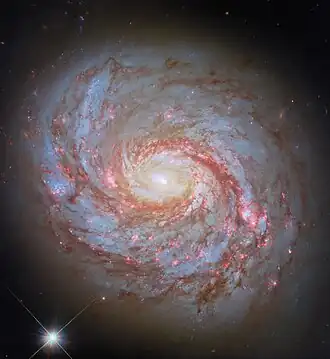 "A close-up of a spiral galaxy, seen face-on. Its center is a bright glowing From the sides of the galaxy’s core emerge spiral arms which wind through the round disc of the galaxy, filled with shining pink spots where stars are forming and more dark-red dust. Some faint stars can be seen around the galaxy, as well as a particularly bright star in the lower left of the image."