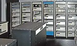 A room with a black box in the foreground and six control cabinets with space for five to six racks each. Most, but not all, of the cabinets are filled with white boxes.