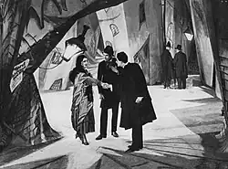 A black-and-white film still.  Three figures stand facing each other on a heavily stylized street set.