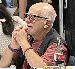 Claremont seated and grinning with his arms crossed