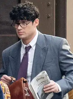 Clark Kent actor David Corenswet during filming in 2024, wearing glasses and a suit while carrying a newspaper and a briefcase