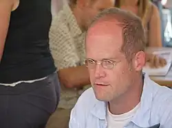 A color photograph of a seated, middle-aged man with a receding hairline. He is wearing spectacles, a white undershirt and a light blue collared shirt. He looks left past the camera.