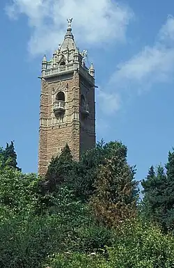 An ornate brick tower surrounded by trees. The tower has balconies and is surmounted by a pitched roof with an ornate figure at the apex.