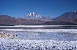 Mountains, including a snow-covered one in the middle, rise above a white surface with a lake
