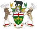 A central shield with the upper part showing the red cross of St. George and the lower part showing three golden maple leaves on a green background. There is a black bear on top of a knight's helmet above the shield with a moose to the left and a Canadian deer to the right. The province's motto "Ut incepit Fidelis sic permanet", Latin for "Loyal she began, loyal she remains" is written below the crest.