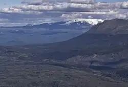A glaciated, flat-topped, gently-sloping mountain shadowed by clouds with a much lower barren mountain in the right foreground.