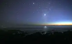 A photograph of the night sky taken from the seashore. Many glimmers of sunlight is on the horizon. There are many stars visible. Venus is at the center, much brighter than any of the stars, and its light can be seen reflected in the ocean.