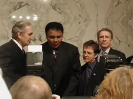 Michael J. Fox and Muhammad Ali are seen speaking to reporters, with the marble walls of the U.S. Senate behind them