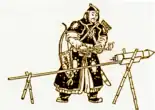 A man in black armor standing in front of a rocket, attached to a stick, with the stick being held up by two X-shaped wooden brackets.