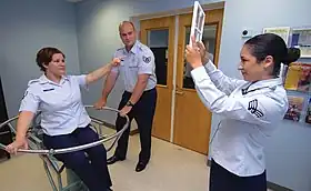 A test person is sitting in a swivel chair. A second person has just stopped the rotation of the chair. The disoriented test person points at a sign held up by a third person.