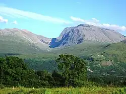 A picture of Ben Nevis on a clear day. A clear blue sky, a large mountain and a very green forest.