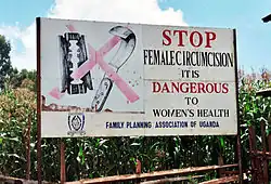 Roadside billboard saying "Stop female circumcision. It's dangerous to Women's health. Family Planning Association of Uganda." Displaying a crossed out razorblade and knife on the left.