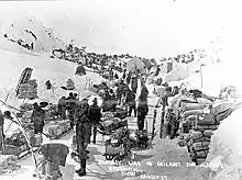 Peak of Chilkoot Pass in March-April 1898. Men wearing winter clothes with their supplies in the snow all of it surrounded by hill-sides.