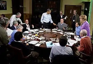 Liderii mondiali la Summit-ul G8 din 2012. (În sensul acelor de ceasornic) Obama (în picioare), Cameron, Medvedev, Merkel, Van Rompuy, Barroso, Noda, Monti, Harper și Hollande.