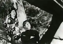 Une jeune femme (Kinuyo Tanaka) en kimino et pieds nus est assise sur la branche d’un arbre. Un étudiant (Den Obinata) les bras croisés a le dos appuyé sur le tronc de l’arbre (image en noir et blanc).