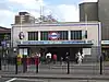 A grey-bricked building with a rectangular, dark blue sign reading "MILE END STATION" in white letters all under a light blue sky with white clouds