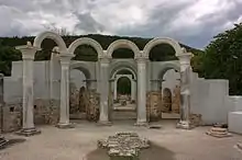 Cinci coloane de marmură lângă pereţii unei clădiri parţial conservate. O poartă şi un monument mic sunt pe fundal.