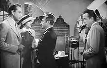 Black-and-white film screenshot of several people in a nightclub. A man on the far left is wearing a suit and has a woman standing next to him wearing a hat and dress. A man at the center, wearing a military uniform and hat, is looking at the man on the left. A man on the far right is wearing a suit and looking to the other people.