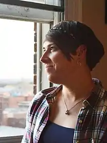 O femeie transgender care privește fericită pe o fereastră. Femeia are părul scurt și poartă o cămașa în carouri. Pe fereastră se vede peisajul unui oraș.