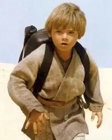 Un băiat blond cu haină gri și purtând la spate un rucsac merge prin deșert.