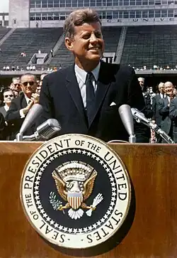 Kennedy vorbind din fața unui podium. Stadionul Rice University se vede în spate.