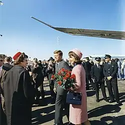 Kennedy și prima doamnă, îmbrăcată într-un costum roz și ținând un buchet de flori, pleacă de la Air Force One și salută publicul.