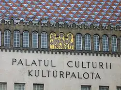 Inscripția bilingvă de pe fațada Palatului Culturii sub mozaicul realizat de Aladár Körösfői-Kriesch pe tema „Patrona Hungariae” (Hódolat Hungáriának)
