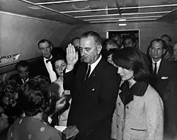 Lyndon B. Johnson cu mâna ridicată deasupra unei biblii în timp ce depune jurământul ca președinte. Jacqueline Kennedy, încă în hainele pătate de sânge (petele nu se văd), privește.