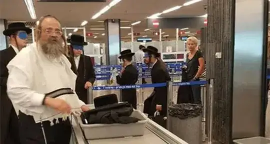 Hasidic men, wearing blindfolds at an airport, being guided by an elder
